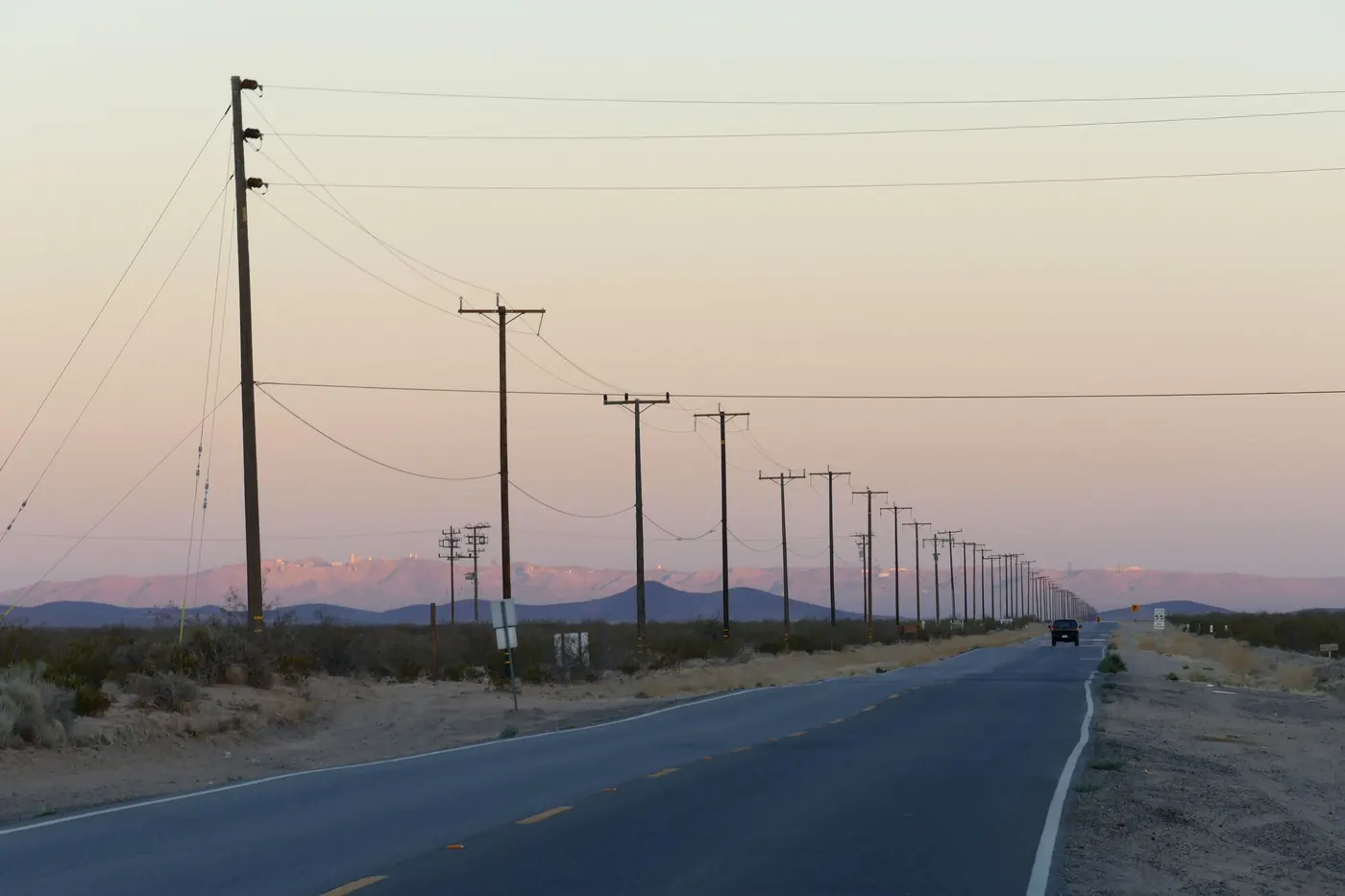 A two-lane road running straight into the distance at dusk, lined by a long row of wooden power poles and lines, with one car on it and pink-lit mountains on the horizon.
