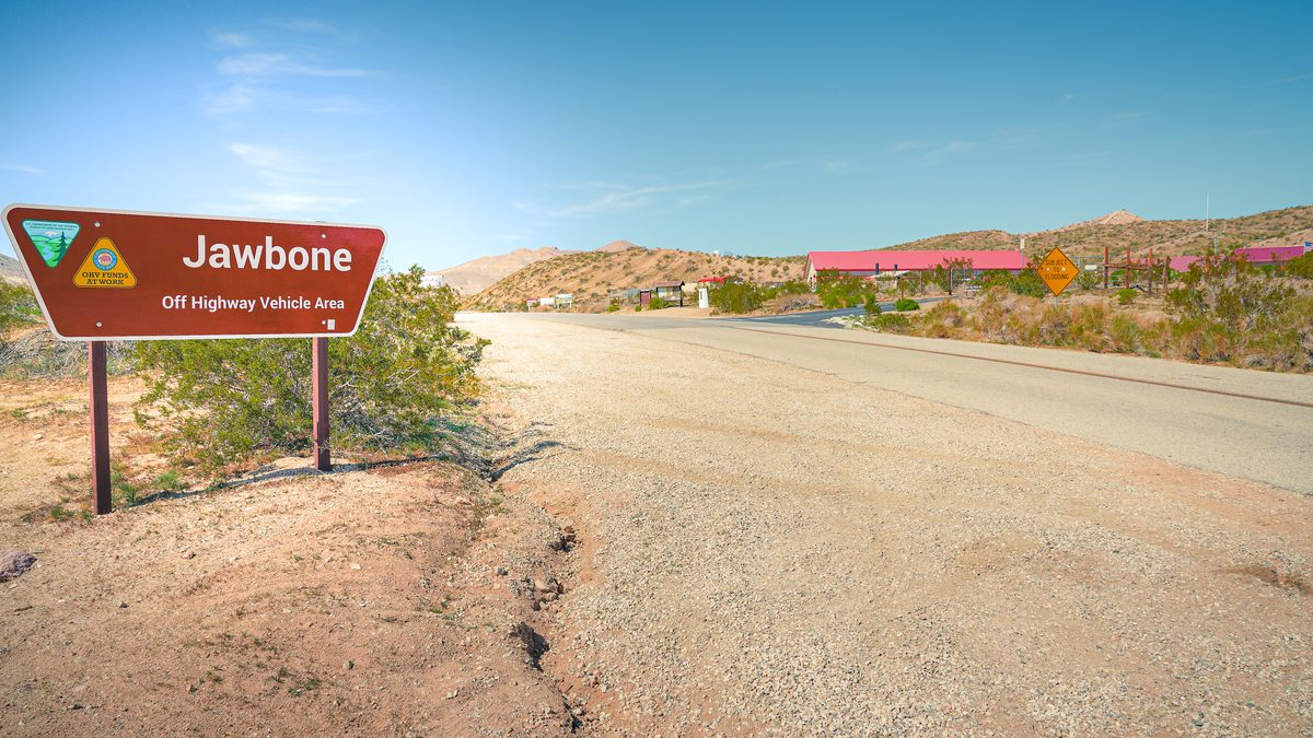 A brown Bureau of Land Management sign reading Jawbone Off Highway Vehicle Area beside a paved road, with low buildings with red roofs and desert hills behind.