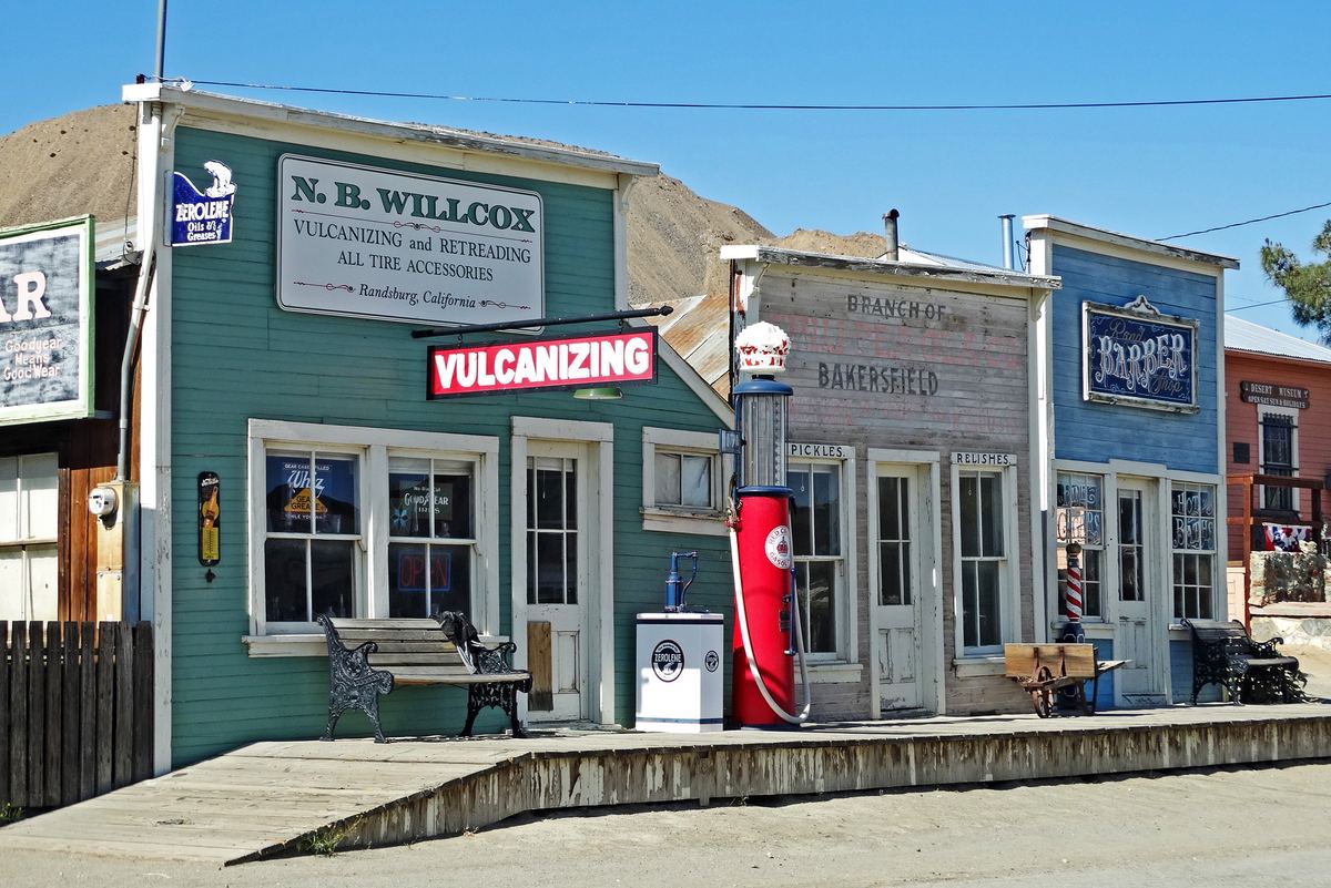 Old wooden false-front shops along a raised boardwalk, painted green and blue, with a vintage red gas pump, a bench and old signs for vulcanizing and a barber.
