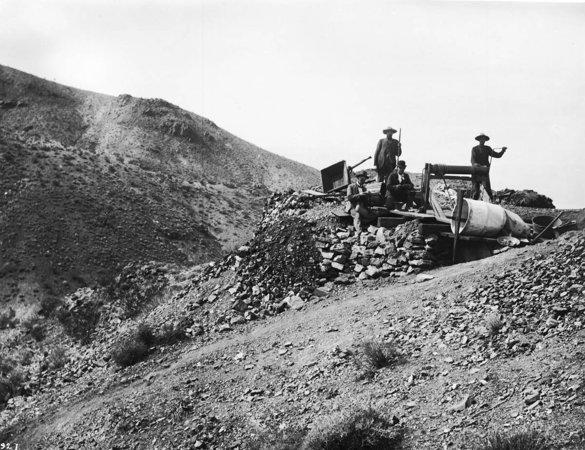 Black-and-white photograph of four men, two standing and two sitting, at a wooden windlass over a prospecting shaft on a rocky hillside strewn with mine waste.