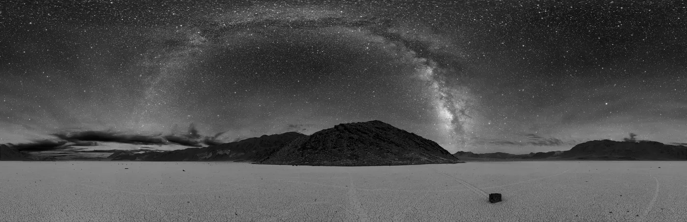 Black-and-white panorama of the Milky Way arching across a starry night sky over a dark hill and a flat, pale playa in Death Valley.