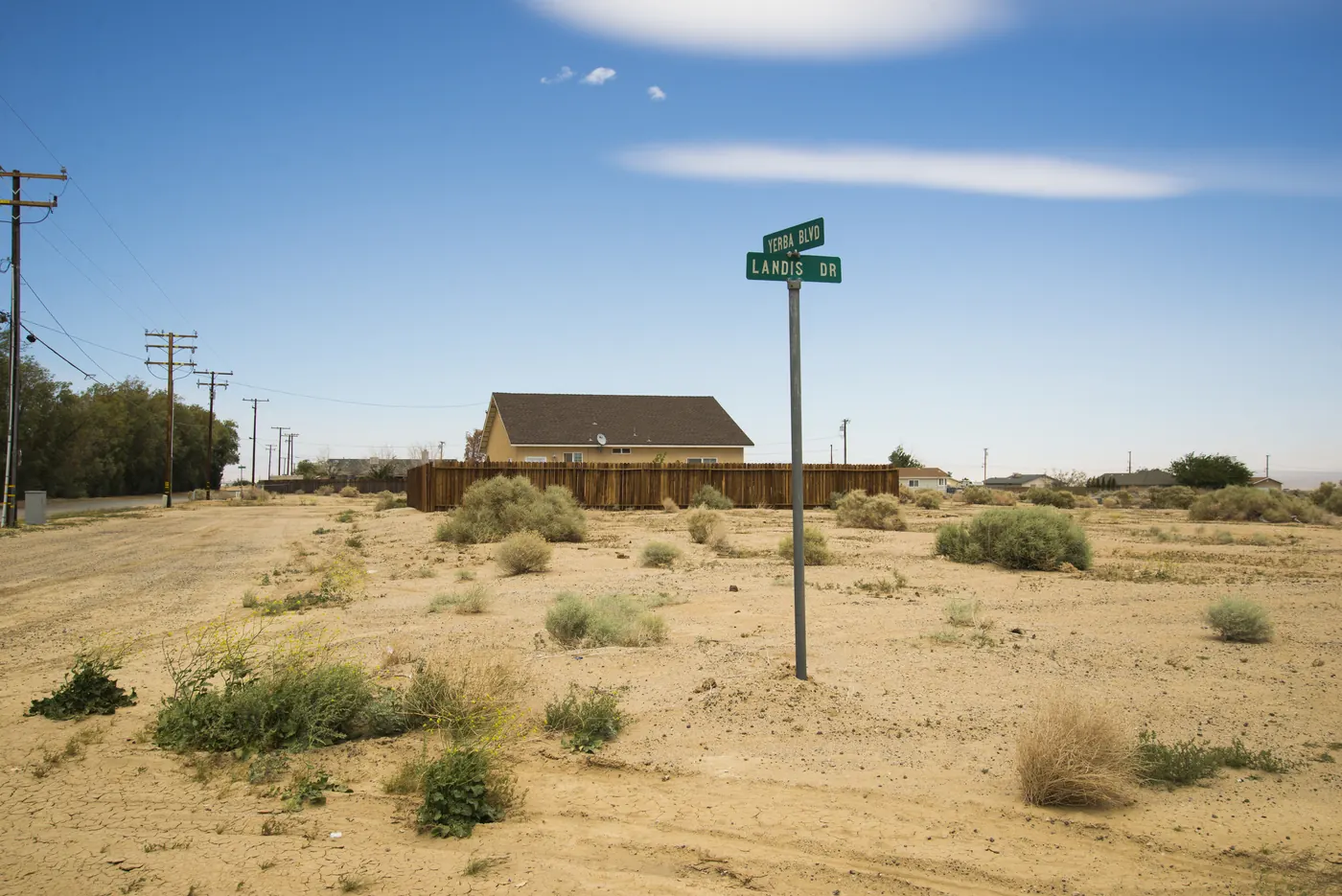 A street sign for Yerba Boulevard and Landis Drive on an unpaved desert corner, with a single-storey house behind a wooden fence and power lines running along the street.