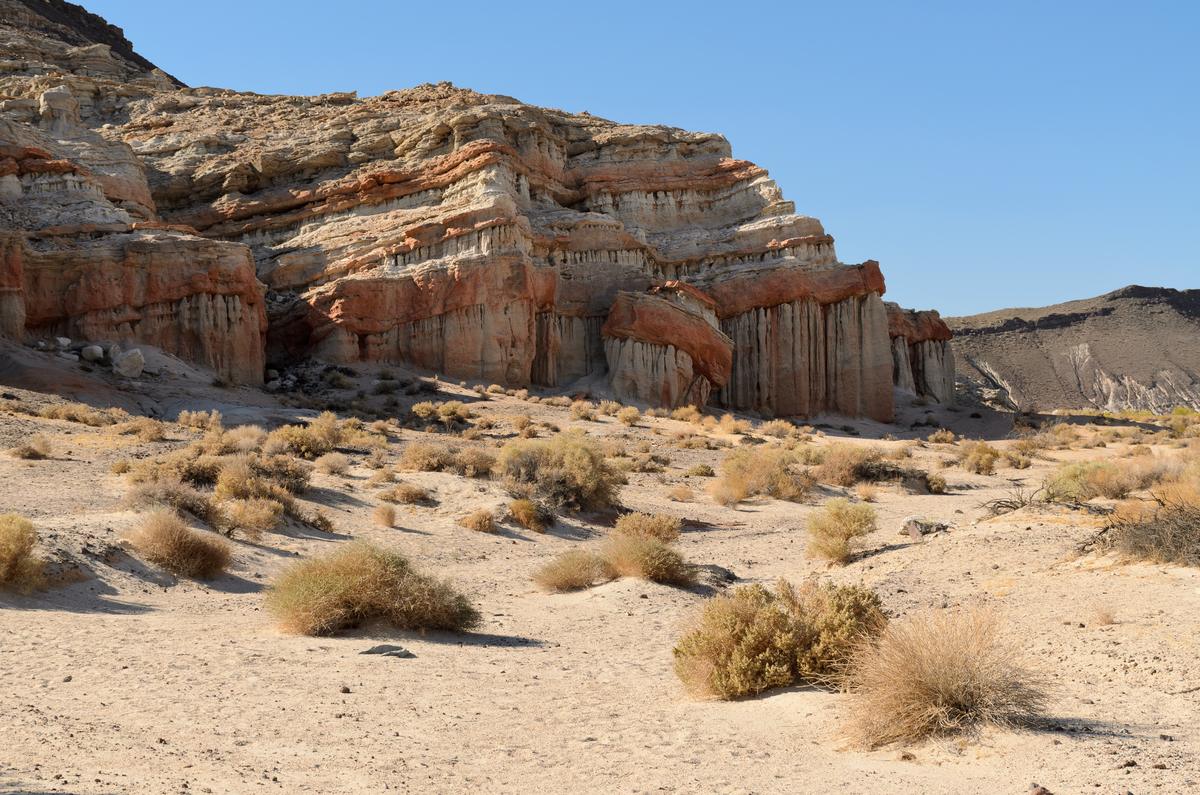 Eroded cliffs layered in pink, red and white bands above a sandy desert floor dotted with dry bunchgrass, under a clear blue sky.