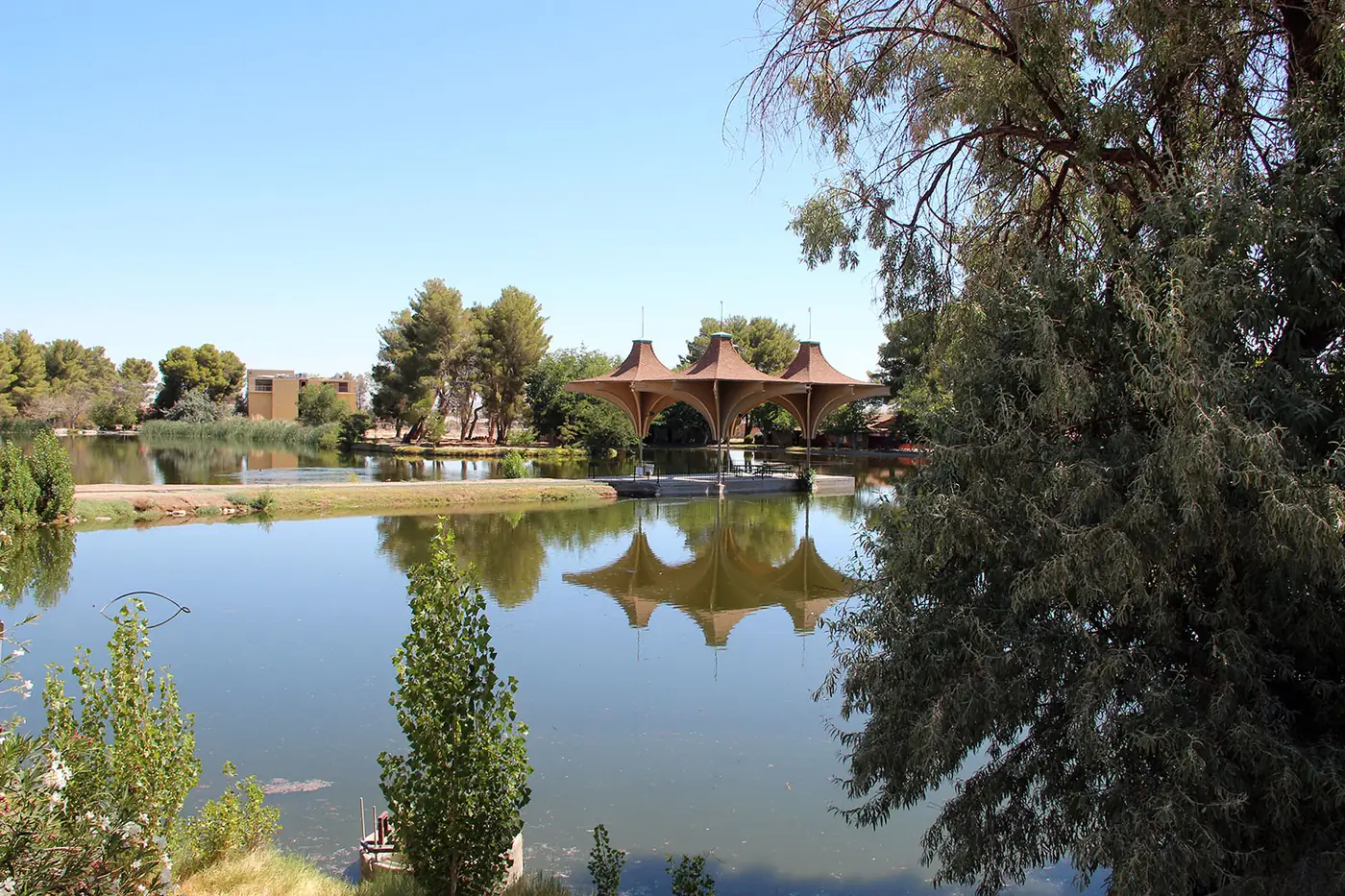 Still water in Central Park's lake reflecting a pavilion with three peaked canvas roofs, pine trees and a tan building on the far shore, framed by a large tree on the right.